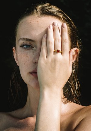 Goldring mit einem herzförmigen roten Edelstein an einer Hand, die die Hälfte eines Gesichts bedeckt, mit heller Haut und nassen, welligen Haaren.
