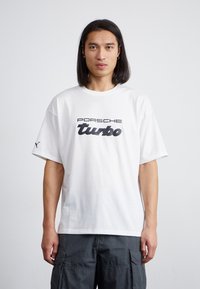 Balts kokvilnas T-krekls ar melnu "Porsche Turbo" grafiku priekšpusē. Brīvs piegriezums ar īsām piedurknēm, vienkārša aizmugure un Puma logo uz piedurknes.