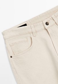 Nahaufnahme von beigen Jeanshosen, die die vordere Tasche, die Gürtelschlaufen und die Metallknopfdetails am Bund zeigen.