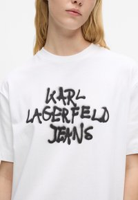 Bijela pamučna majica s crnim logotipom u spreju koji glasi "KARL LAGERFELD JEANS." Okrugli izrez i opušteni kroj.