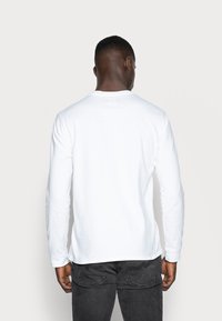 Long-sleeve white T-shirt in soft fabric with a crew neckline, worn with dark gray jeans, showing a smooth back and fitted silhouette.