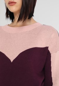 Svetr s pulovrovým střihem v růžové a tmavě fialové barvě s designem ve tvaru srdce. Vyroben z jemné pleteniny, má kulatý výstřih a žebrované lemy.