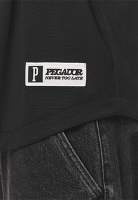 Czarny teksturowany T-shirt z białą prostokątną etykietą z napisem "PEGADOR" i "NEVER TOO LATE", w połączeniu z ciemnymi dżinsami.