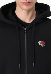 Sudadera con cremallera negra hecha de un tejido suave, que presenta un diseño de rosa bordada en la zona del pecho izquierdo y cordones ajustables en la capucha.