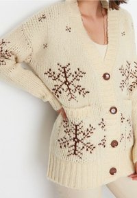 Cremefarbene Strickjacke mit braunem Schneeflockenmuster, gerippter Struktur, großen Knöpfen und Taschen, mit V-Ausschnitt und oversized Passform.