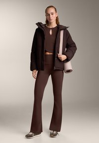 Vrouw draagt bruine atletische uitlopende leggings, cropped top, zwarte pufferjas, sneakers, met een opgerolde yogamat over haar schouder.