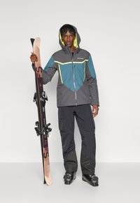 Casaco de esqui cinza com detalhes em azul turquesa e amarelo, combinado com calças de neve pretas e botas de esqui, segurando esquis de madeira com fixações pretas.