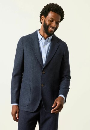 Mann lächelnd trägt eine dunkelblaue Anzugsjacke und passende Hose über einem hellblauen Hemd mit Knöpfen vor einem einfarbigen Hintergrund.