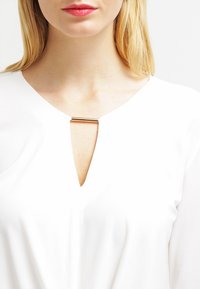 Blusa bianca con scollatura a V e accentuazione dorata. Il tessuto ha una consistenza liscia e una vestibilità leggermente ampia.