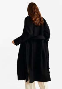 Černý dlouhý kabát s páskem, vyrobený z měkké tkaniny, s nadměrnými rukávy a bočními kapsami, zobrazený zezadu na neutrálním pozadí.