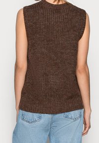 Brauner gestrickter ärmelloser Pullover mit Rippstruktur und hohem Kragen, mit einem taillierten unteren Saum, getragen über hellblauen Jeans.