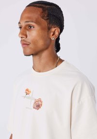 Camiseta de algodón color crema con un gráfico de dos rosas y un diseño de cruz, cuello redondo y corte relajado.