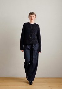 Μαύρο ριγωτό καπιτονέ με κουμπιά, συνδυασμένο με σκούρες ραβδωτές παντελόνες με φαρδύ πόδι; το μοντέλο στέκεται σε ξύλινο δάπεδο απέναντι από έναν απλό τοίχο.