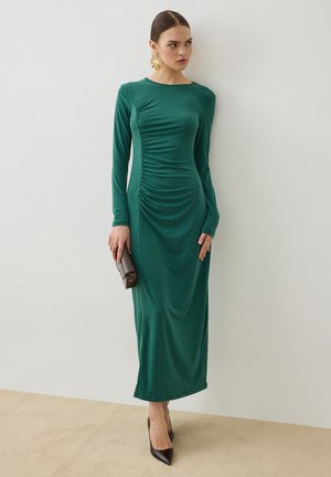Femme en robe verte ajustée à manches longues tenant une pochette sombre, portant des talons noirs et des boucles d'oreilles dorées voyantes, debout contre un mur uni.