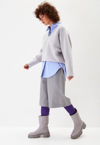 Suéter gris recortado sobre una camisa de rayas azules claras, pantalones cortos grises, leggings morados y botas hasta la rodilla grises con suelas gruesas.