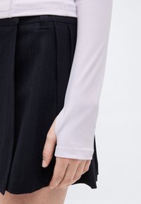 Χέρι ξεκουρασμένο στο πλάι, φορώντας λευκό μακρυμάνικο τοπ με άνοιγμα για τον αντίχειρα και μαύρη πλισέ φούστα με λεπτές ρίγες.