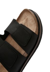 Sandal nera con due fasce larghe, realizzata in pelle liscia, e un plantare beige testurizzato con un leggero arco. Suola in gomma con accenti marroni.