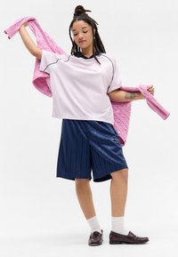 Ανοιχτό ροζ υπερμεγέθη πουκάμισο με μαύρες λεπτομέρειες, συνδυασμένο με σκούρα μπλε κοντά παντελόνια με ρίγες και ροζ πουλόβερ με πλεξούδα, φορεμένο από ένα άτομο.