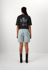 Svart grafisk t-shirt med en design som har text och logotyper, kombinerad med ljusblå texturerade shorts och svarta knähöga stövlar.