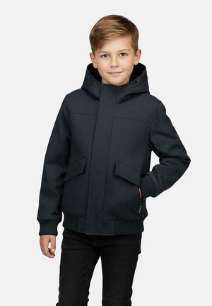 Boy wearing a black hooded jacket with front pockets, standing with one hand in pocket and a slight smile against a plain background.