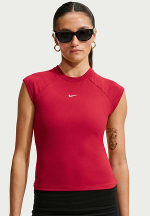 Frau mit dunklen Haaren im Pferdeschwanz, trägt schwarze Sonnenbrille, goldene Creolen-Ohrringe, rotes ärmelloses Nike-Top und schwarze Hose, mit einem Tattoo am Arm.