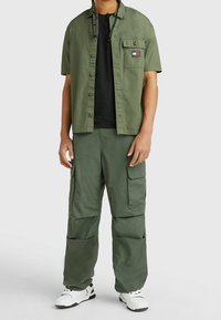 Hombre vestido con pantalones cargo verde oliva y una camisa de manga corta sobre una camiseta negra, combinados con zapatillas blancas y negras, de pie contra un fondo liso.