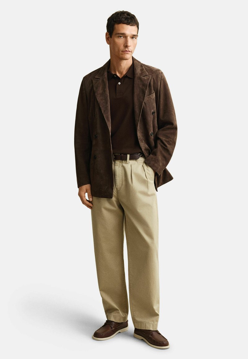 Man draagt een bruine suède blazer, donkerbruine polo, beige geplooide broek met bruine riem en bruine mocassins, staand tegen een witte achtergrond.