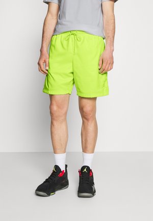 Mann trägt neongrüne Sportshorts, graues Shirt, weiße Socken und schwarze Sneakers mit roten und gelben Akzenten und steht auf grauem Boden.