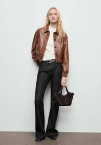 Chaqueta bomber de cuero marrón con botones negros, combinada con un suéter ligero y pantalones oscuros de campana. Sosteniendo un pequeño bolso oscuro.