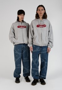 Gray hooded sweatshirts with red logos, paired with blue wide-leg jeans featuring a chain and knuckle pattern, worn with black shoes.