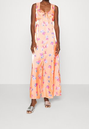 Mouwloze maxi-jurk van perzik satijn met roze en paarse bloemmotieven, een strikdetail aan de voorkant, gedragen met zilveren open-toe slide sandalen.