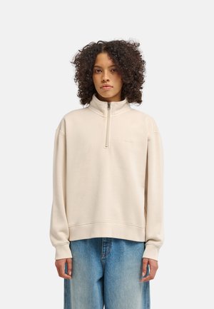 Giovane con capelli ricci indossa una felpa beige con mezza cerniera e jeans blu larghi, in piedi davanti a uno sfondo chiaro e uniforme.