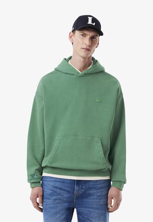 Sudadera verde de algodón con bolsillo canguro y puños acanalados. Presenta un pequeño logo bordado en el pecho. Combinada con jeans de mezclilla azul.