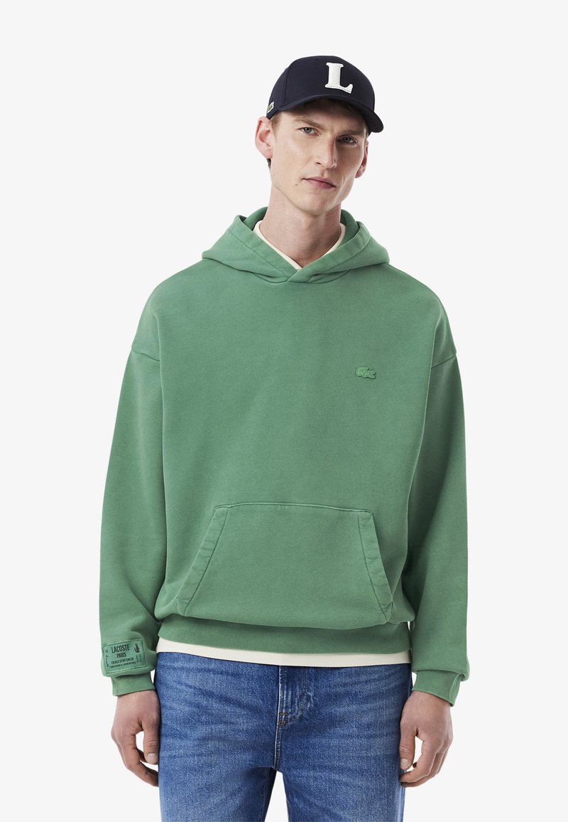 Grüne Baumwollkapuzenjacke mit Kängurutasche und gerippten Bündchen. Verfügt über ein kleines gesticktes Logo auf der Brust. Kombiniert mit blauen Jeans.