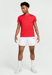 Joven vestido con una camiseta roja de Nike, pantalones cortos blancos de Nike, calcetines blancos de Nike y zapatillas blancas de Nike, de pie contra un fondo liso.