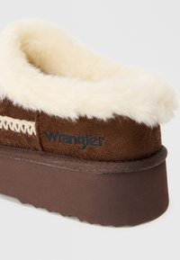 Brūns čības ar pūkainu krēmkrāsas odere, teksturētu zamšu augšdaļu un biezu, izturīgu brūnu zoli ar rievotām malām. Wrangler logo redzams.