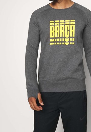 Mann trägt ein dunkelgraues Langarmshirt mit gelbem "BARÇA"-Schriftzug und schwarze Sporthose mit sichtbarem Nike-Logo am linken Bein.