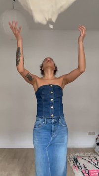 Asmuo vilki džinso be petnešėlių viršutinę dalį ir sijoną, mėto lengvą audinį virš galvos, stovėdamas minimalistiškai įrengtame kambaryje su raštuotu kilimu ant grindų.