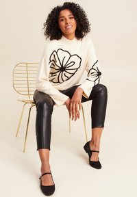 Maglione crema con design floreale nero, abbinato a pantaloni in pelle nera e scarpe basse nere. Sedia dorata sullo sfondo, sfondo chiaro e semplice.