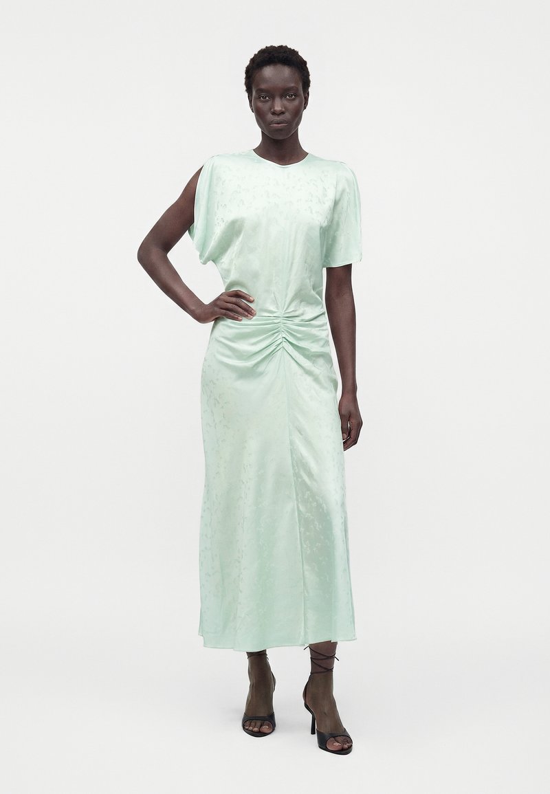 Femme portant une robe midi en satin vert clair avec une taille froncée et des manches courtes, debout avec une main sur la hanche et des sandales noires à talons.