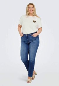 T-shirt branca com um patch de coração de leopardo, combinada com jeans skinny azul escuro e sandálias de tacão bege. Em um fundo neutro.