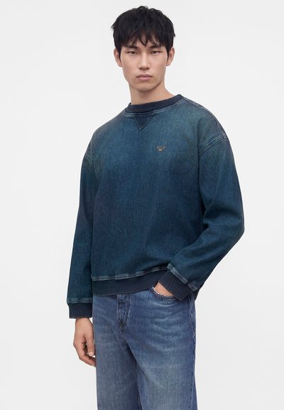Jeune homme portant un sweat en denim bleu foncé et un jean bleu, une main dans la poche, debout devant un fond clair uni.