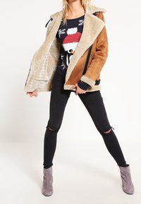 Modellen poserar självsäkert, iklädd en brun och beige jacka över en festlig pingvintröja och svarta jeans med hål, kompletterad av grå stövletter. 