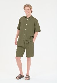 Oliivinvihreä lyhythihainen paita, jossa on napit ja yhteensopivat lyhyet housut. Materiaali vaikuttaa kevyeltä; päällä ruskeat sandaalit.