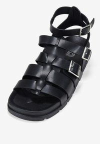 Sandalia negra con un diseño de tira trenzada, hebillas ajustables y una suela texturizada. Hecha de cuero sintético con un diseño plano.