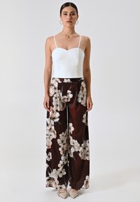 Witte aansluitende bodysuit met dunne bandjes in combinatie met wijde broekspijpen met een donkerbruine achtergrond en grote lichte bloemmotieven.