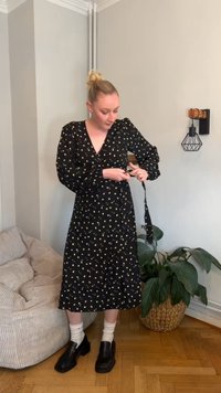 Svart omlottklänning med blommönster, långa ärmar och djup V-ringning. Modellen kombinerar den med svarta slip-on skor och vita strumpor.