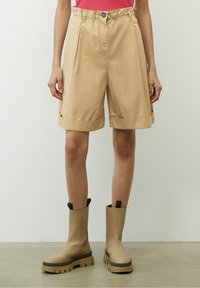 Persona vestita con pantaloni beige a vita alta, plissettati e con orli risvoltati, abbinati a stivali chunky a suola spessa color beige, che si trovano su un pavimento di cemento.