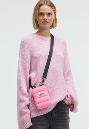 Pinke, gepolsterte Umhängetasche mit schwarzem, verstellbarem Riemen, ausgestattet mit einer Fronttasche und Reißverschluss. Getragen über einem hellrosa Pullover.