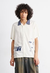 Camicia a maniche corte color crema con colletto blu navy. Presenta grafiche stampate in blu, tra cui disegni astratti e testo, realizzata in tessuto leggero.